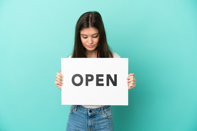 Joven mujer caucásica aislada sobre fondo azul sosteniendo un cartel con texto ABIERTO