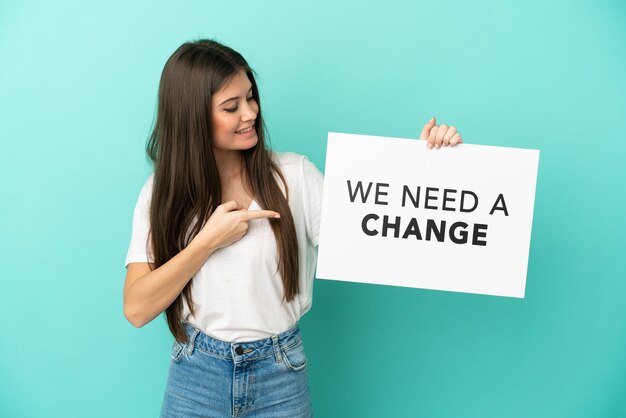 Joven mujer caucásica aislada sobre fondo azul sosteniendo un cartel con el texto Necesitamos un cambio y apuntando