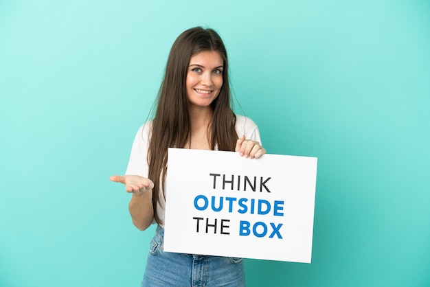 Joven mujer caucásica aislada sobre fondo azul sosteniendo un cartel con texto pensar fuera de la caja haciendo un trato