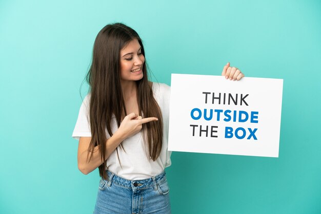 Joven mujer caucásica aislada sobre fondo azul sosteniendo un cartel con el texto Piense fuera de la caja y apuntándolo