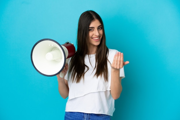 Foto joven mujer caucásica aislada sobre fondo azul sosteniendo un megáfono e invitando a venir con la mano