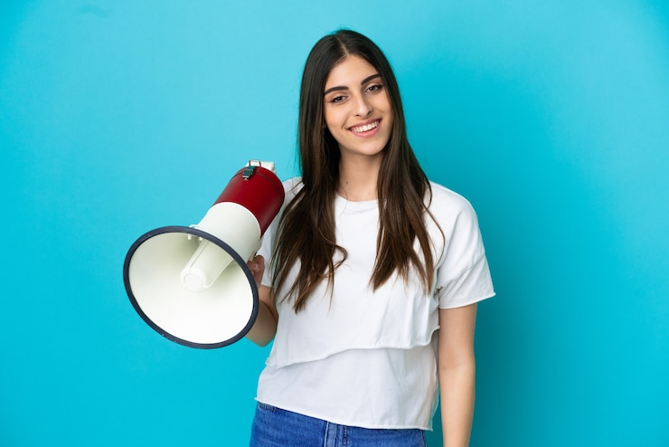 Foto joven mujer caucásica aislada sobre fondo azul sosteniendo un megáfono y sonriendo mucho
