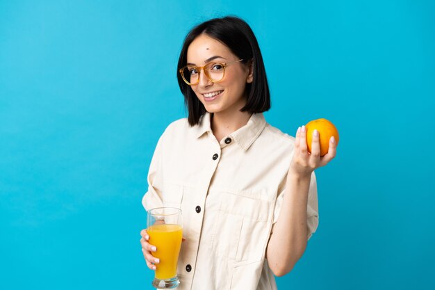 Joven mujer caucásica aislada sobre fondo azul sosteniendo una naranja y un jugo de naranja