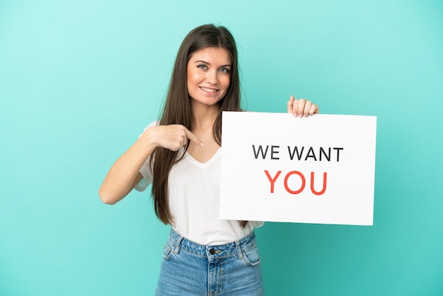 Joven mujer caucásica aislada sobre fondo azul sosteniendo We Want You board y apuntando