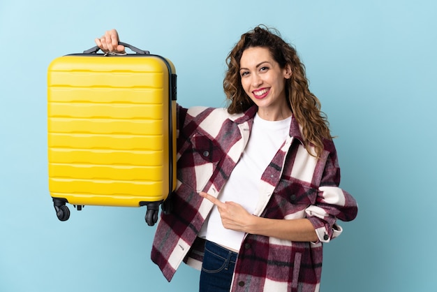 Joven mujer caucásica aislada sobre fondo azul en vacaciones con maleta de viaje