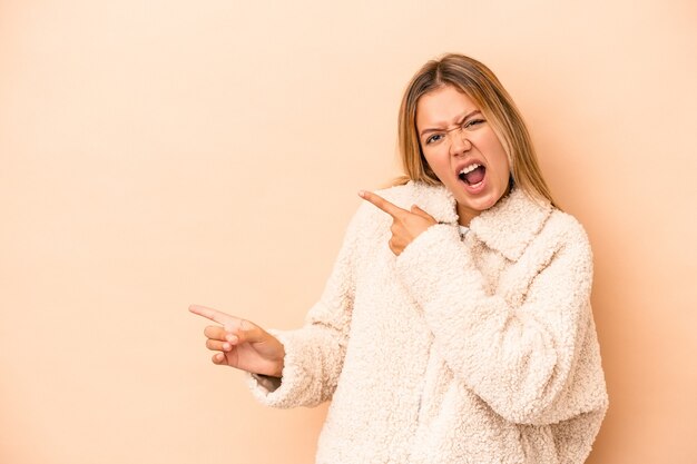 Joven mujer caucásica aislada sobre fondo beige apuntando con los dedos índices a un espacio de copia, expresando entusiasmo y deseo.