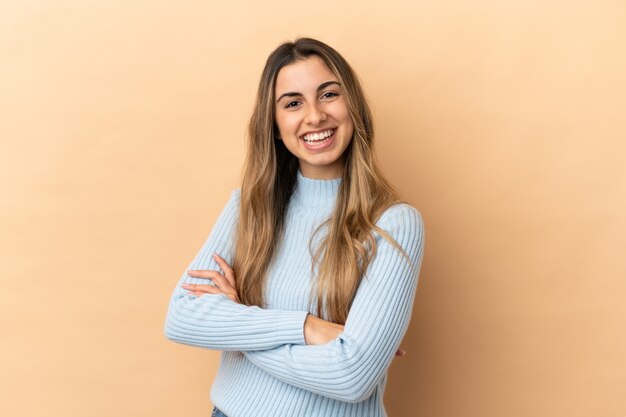 Joven mujer caucásica aislada sobre fondo beige con los brazos cruzados y mirando hacia adelante