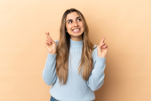 Joven mujer caucásica aislada sobre fondo beige con los dedos cruzando y deseando lo mejor