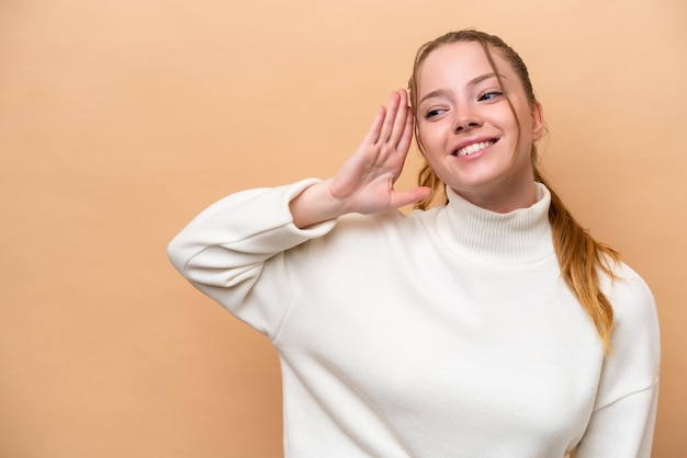 Joven mujer caucásica aislada sobre fondo beige escuchando algo poniendo la mano en la oreja