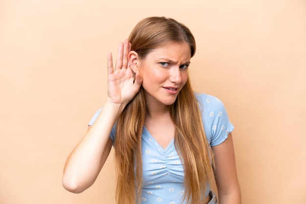 Joven mujer caucásica aislada sobre fondo beige escuchando algo poniendo la mano en la oreja