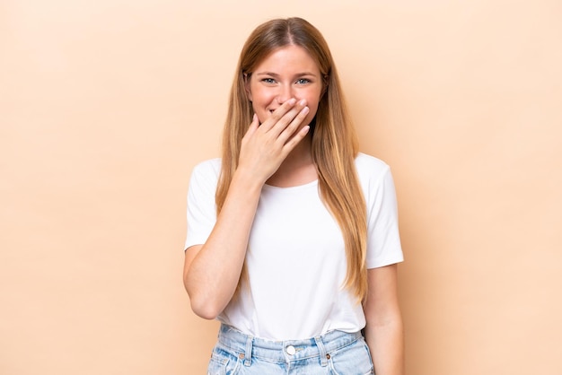 Joven mujer caucásica aislada sobre fondo beige feliz y sonriente cubriendo la boca con la mano