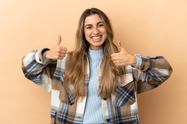 Joven mujer caucásica aislada sobre fondo beige con gesto de pulgar hacia arriba y sonriente