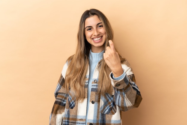 Joven mujer caucásica aislada sobre fondo beige haciendo gesto de venida