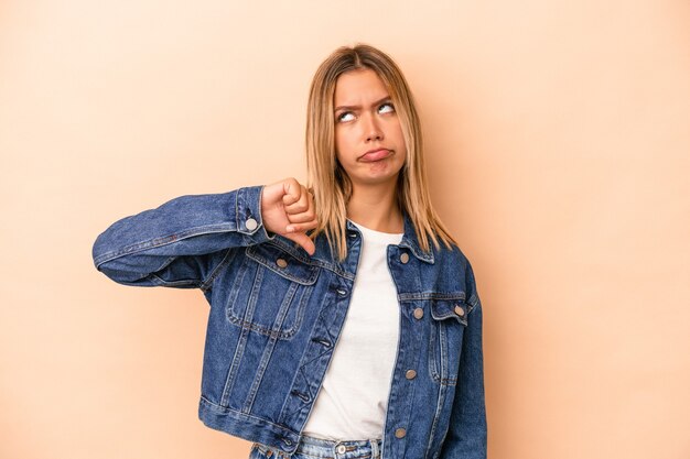 Joven mujer caucásica aislada sobre fondo beige mostrando un gesto de aversión, pulgares hacia abajo. Concepto de desacuerdo.