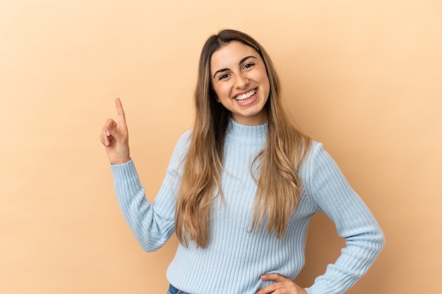 Joven mujer caucásica aislada sobre fondo beige mostrando y levantando un dedo en señal de lo mejor