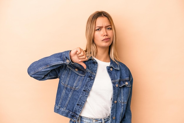 Joven mujer caucásica aislada sobre fondo beige mostrando el pulgar hacia abajo, concepto de decepción.