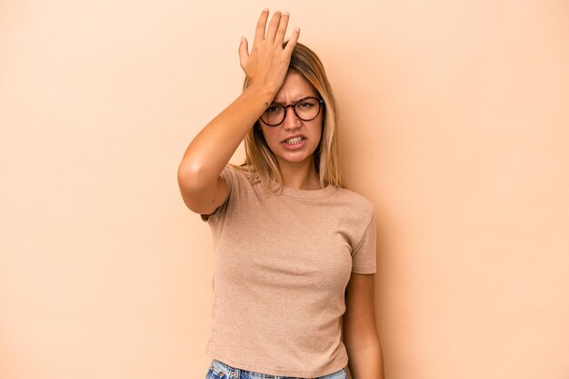 Joven mujer caucásica aislada sobre fondo beige olvidando algo, golpeándose la frente con la palma y cerrando los ojos.