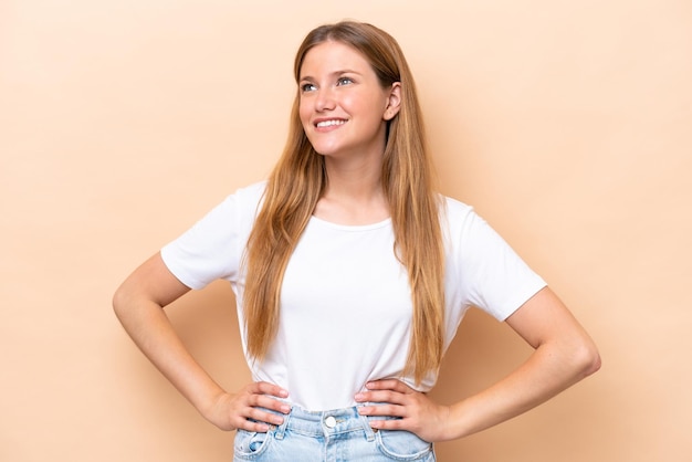 Joven mujer caucásica aislada sobre fondo beige posando con los brazos en la cadera y sonriendo