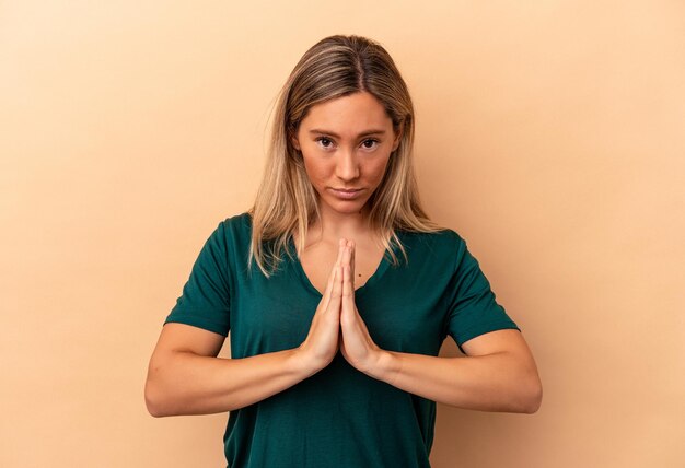 Joven mujer caucásica aislada sobre fondo beige rezando, mostrando devoción, persona religiosa en busca de inspiración divina.