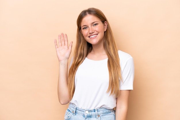 Joven mujer caucásica aislada sobre fondo beige saludando con la mano con expresión feliz