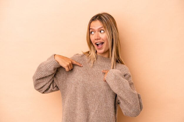 Joven mujer caucásica aislada sobre fondo beige sorprendió señalando con el dedo, sonriendo ampliamente.