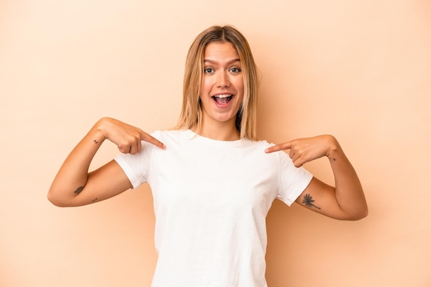 Joven mujer caucásica aislada sobre fondo beige sorprendió señalando con el dedo, sonriendo ampliamente.