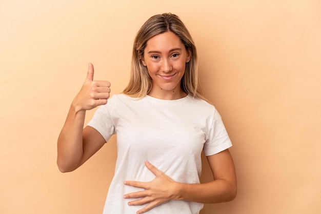 Joven mujer caucásica aislada sobre fondo beige toca la barriga, sonríe suavemente, comiendo y concepto de satisfacción.