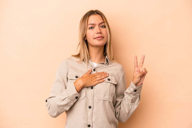 Joven mujer caucásica aislada sobre fondo beige tomando un juramento, poniendo la mano en el pecho.