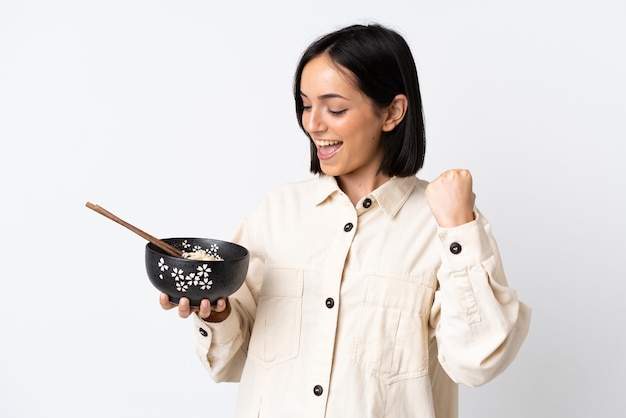 Joven mujer caucásica aislada sobre fondo blanco celebrando una victoria mientras sostiene un plato de fideos con palillos