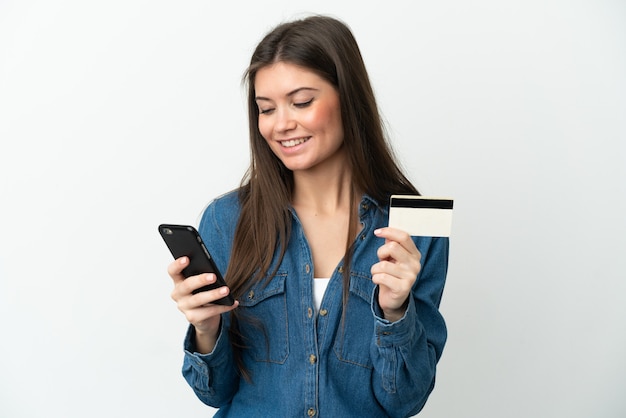 Joven mujer caucásica aislada sobre fondo blanco comprando con el móvil con tarjeta de crédito