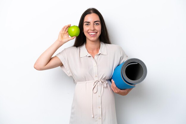 Joven mujer caucásica aislada sobre fondo blanco embarazada sosteniendo una manzana e ir a clases de yoga