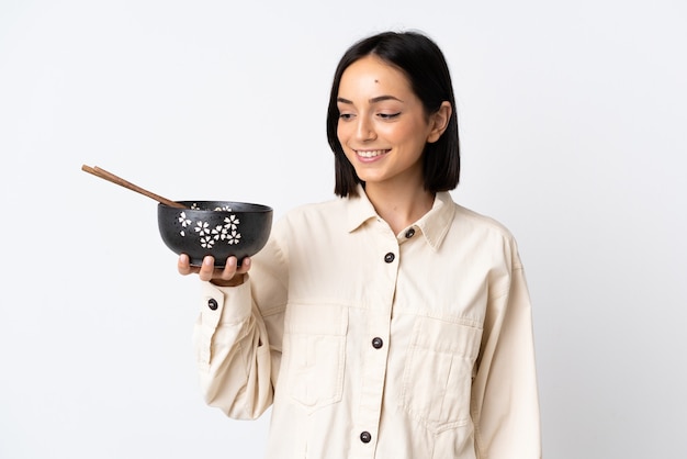 Joven mujer caucásica aislada sobre fondo blanco con expresión feliz mientras sostiene un plato de fideos con palillos