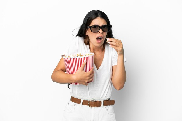 Joven mujer caucásica aislada sobre fondo blanco con gafas 3d y sosteniendo un gran balde de palomitas de maíz