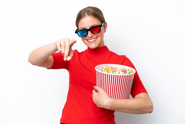 Joven mujer caucásica aislada sobre fondo blanco con gafas 3d y sosteniendo un gran cubo de palomitas de maíz mientras apunta hacia el frente