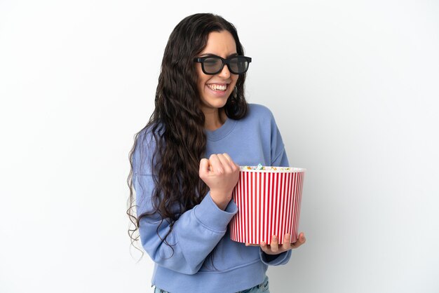 Joven mujer caucásica aislada sobre fondo blanco con gafas 3d y sosteniendo un gran cubo de palomitas de maíz mientras mira de lado