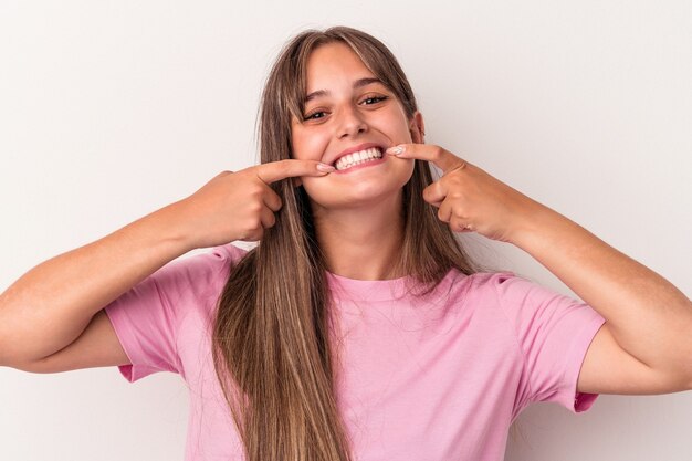 Joven mujer caucásica aislada sobre fondo blanco sonríe, señalando con el dedo a la boca.