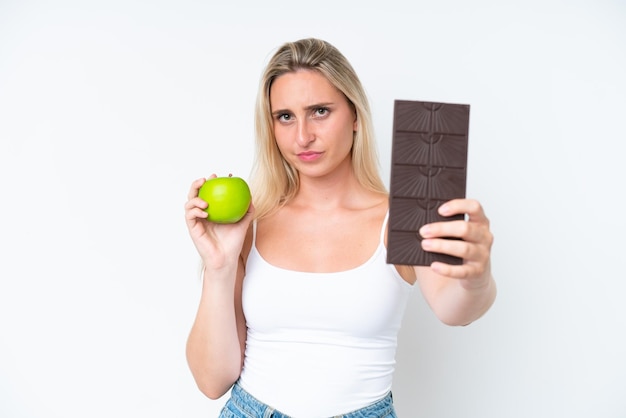 Joven mujer caucásica aislada sobre fondo blanco tomando una tableta de chocolate en una mano y una manzana en la otra