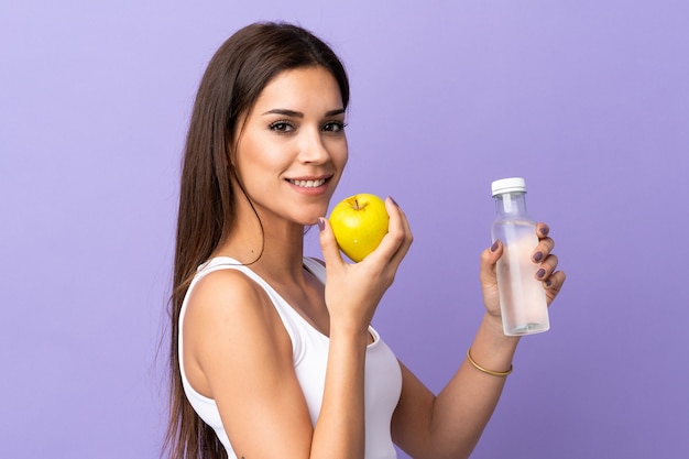 Joven mujer caucásica aislada sobre fondo morado con una manzana y con una botella de agua