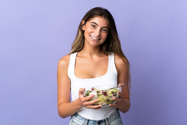 Joven mujer caucásica aislada sobre fondo púrpura sosteniendo un plato de ensalada con expresión feliz