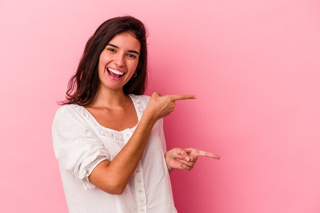 Joven mujer caucásica aislada sobre fondo rosa apuntando con los dedos índices a un espacio de copia, expresando entusiasmo y deseo.