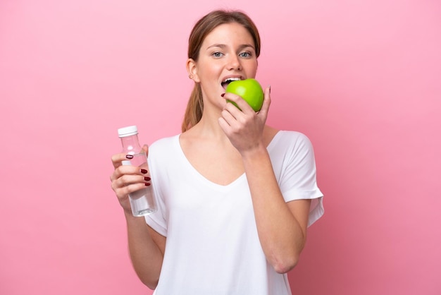 Joven mujer caucásica aislada sobre fondo rosa con una botella de agua y comiendo una manzana