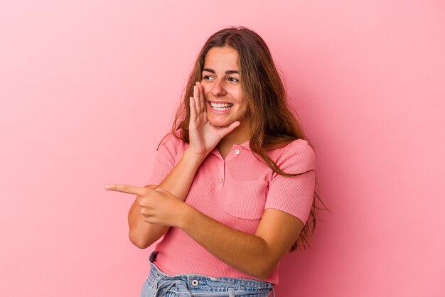 Joven mujer caucásica aislada sobre fondo rosa diciendo un chisme, apuntando al lado informando algo.