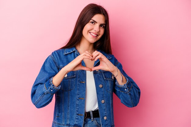 Joven mujer caucásica aislada sobre fondo rosa sonriendo y mostrando una forma de corazón con las manos.