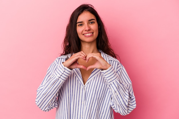 Joven mujer caucásica aislada sobre fondo rosa sonriendo y mostrando una forma de corazón con las manos.