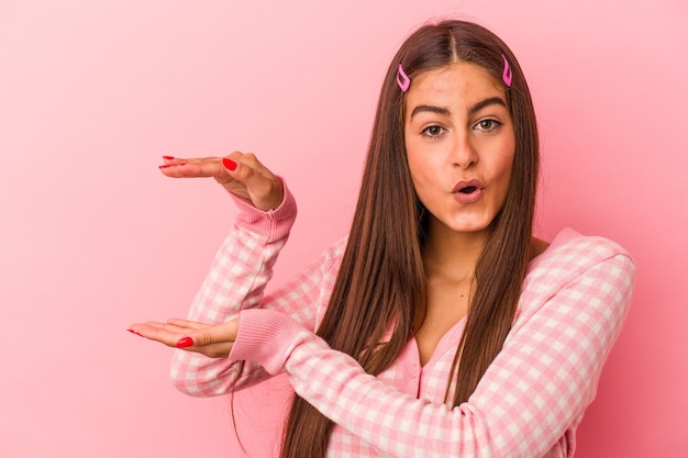 Joven mujer caucásica aislada sobre fondo rosa sorprendido y sorprendido sosteniendo un espacio de copia entre las manos.