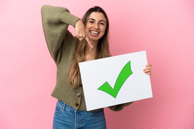 Joven mujer caucásica aislada sobre fondo rosa sosteniendo un cartel con el icono de marca de verificación verde de texto y apuntando