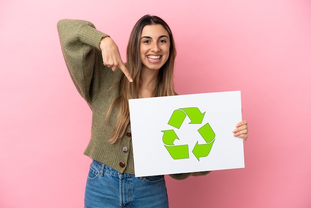 Joven mujer caucásica aislada sobre fondo rosa sosteniendo un cartel con el icono de reciclaje y apuntando hacia él