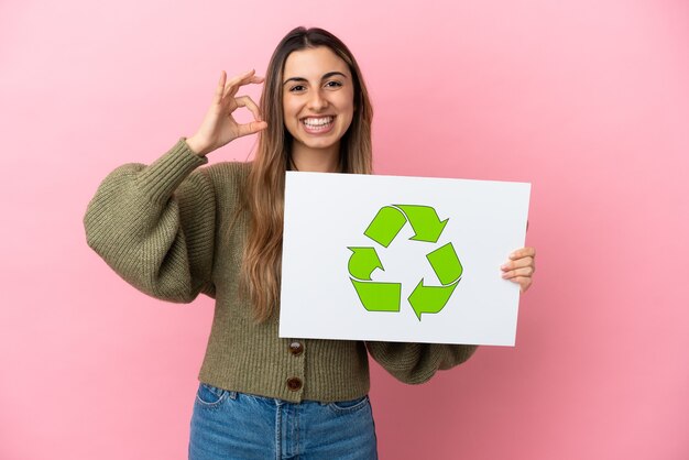 Joven mujer caucásica aislada sobre fondo rosa sosteniendo un cartel con el icono de reciclaje y celebrando una victoria