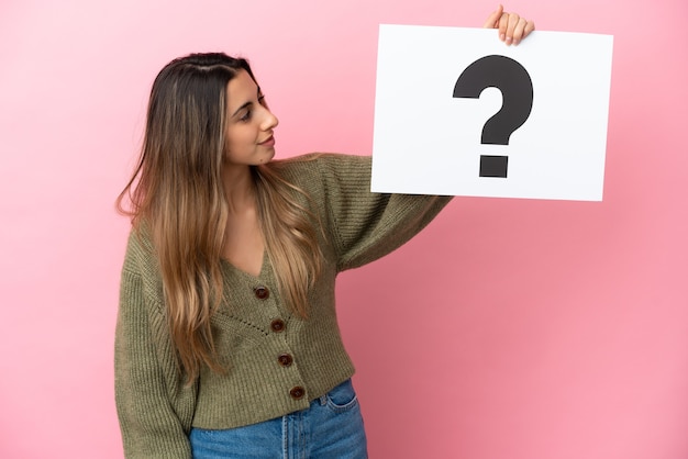 Foto joven mujer caucásica aislada sobre fondo rosa sosteniendo un cartel con el símbolo de interrogación