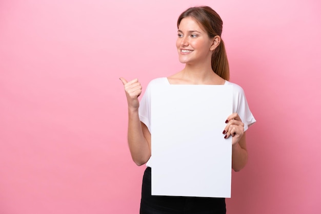 Foto joven mujer caucásica aislada sobre fondo rosa sosteniendo un cartel vacío y apuntando hacia el lado